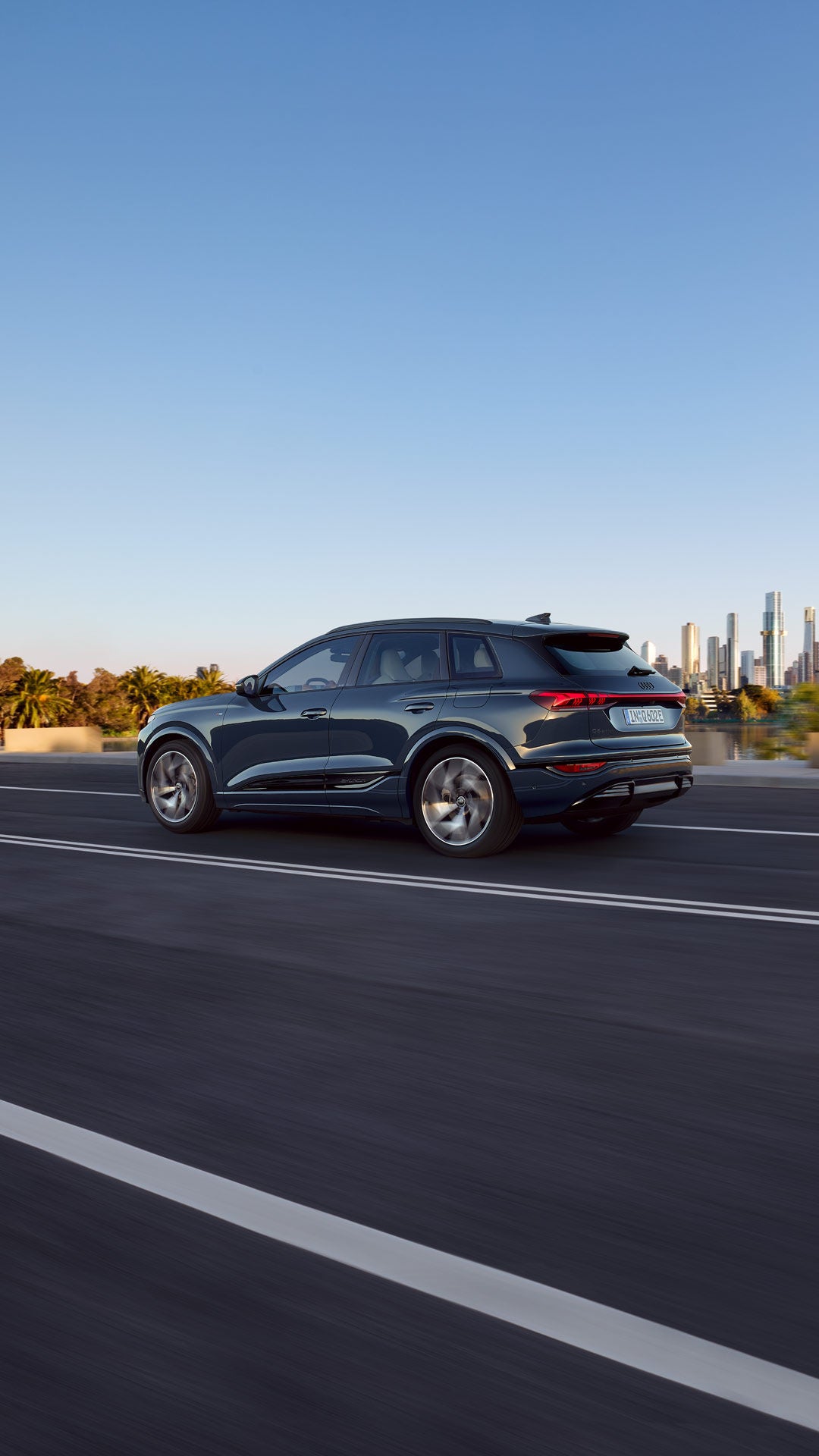 Side profile of the Q6 SUV e-tron driving on a road by a river. The skyline of a city can be seen in the background.