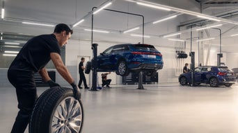Deux Audi bleues sont en train de se faire monter de nouveaux pneus dans un garage. Au premier plan, un employé pousse un pneu neuf sur un chariot en direction des véhicules.