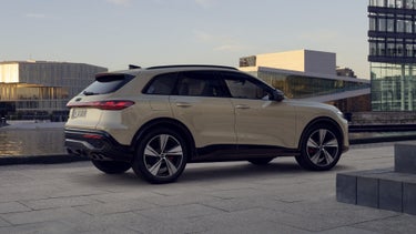 Der Q5 SUV in der edition one mit Kontrasten in Mythosschwarz steht im seitlichen Frontprofil auf einem Parkplatz direkt an der Küste. Dahinter ein futuristisches Haus.