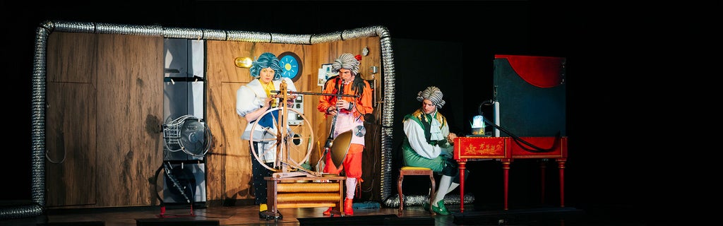 Drei kostümierte Darsteller auf einer Theaterbühne mit buntem Bühnenbild, Spinnrad und rotem Cembalo.