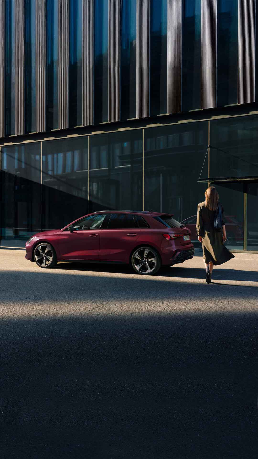 Frau geht auf einen roten Audi A3 Sportback zu, der am Straßenrand vor einem modernen Gebäude steht.