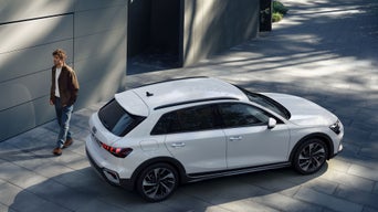 Mann steht vor modernem Gebäude neben geparktem weißen Audi SUV, Ansicht leicht erhöht bei Tageslicht.