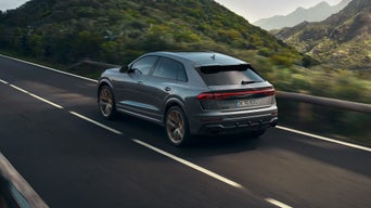 Grauer Audi RS Q8 SUV fährt auf einer kurvigen Straße durch grüne Berglandschaft.