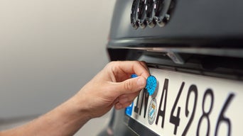 Eine Hand bringt eine blaue Plakette auf einem deutschen Nummernschild an einem schwarzen Audi an.