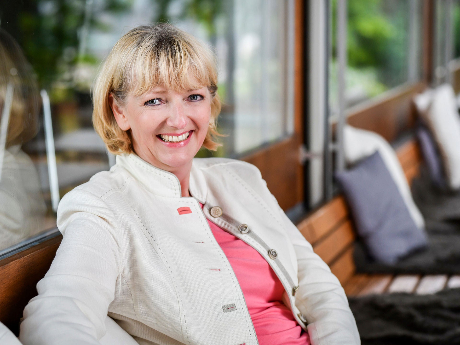 Porträt einer Frau (Gudrun Sander) mit hellem Blazer und rosa Oberteil, die entspannt lächelt.