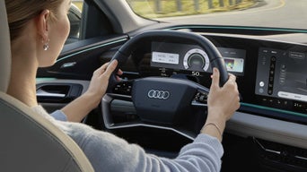 Cockpit of the Q6 SUV e-tron from the rear seat perspective. A young woman sits in the driver's seat, holding the steering wheel with both hands. Through the windscreen, you can see a road surrounded by nature.