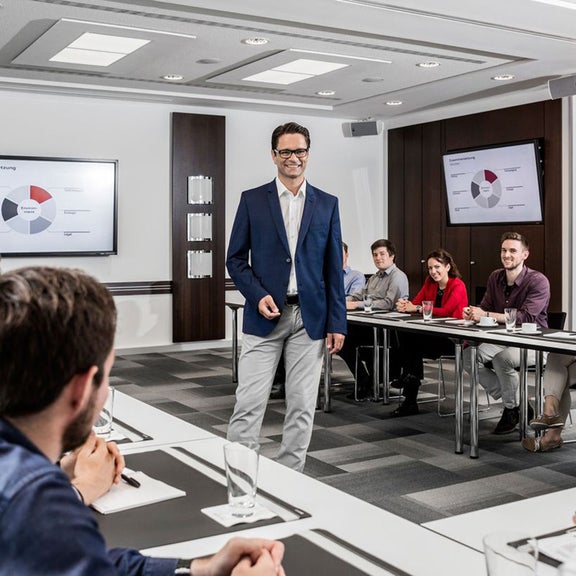 Präsentation mit Diagramm in modernem Konferenzraum, mehrere Personen hören aufmerksam einem Mann mit Brille in Anzug zu. 