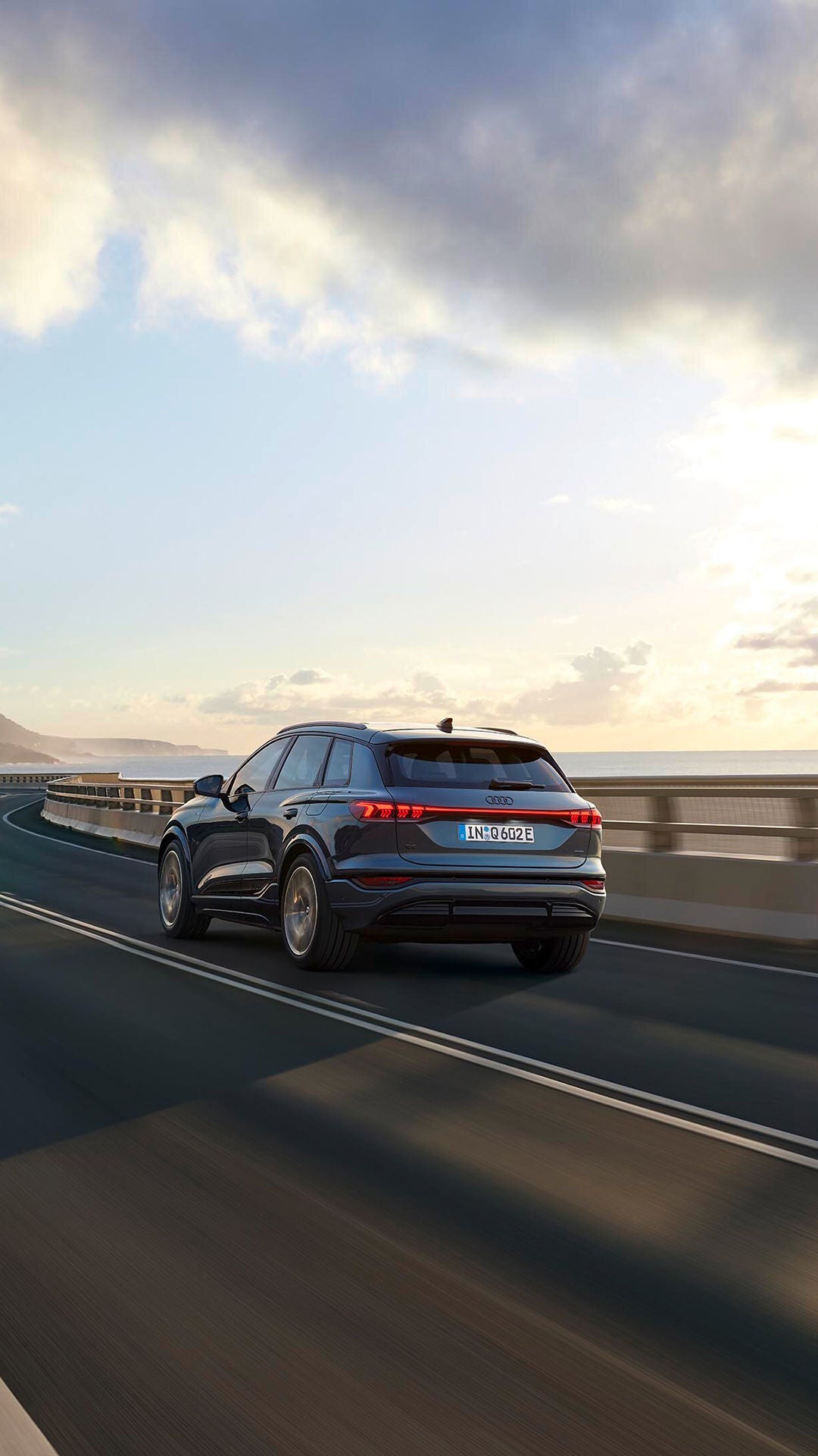 Ein blauer Audi SUV fährt bei Sonnenuntergang auf einer Küstenstraße mit Meerblick und Bergen im Hintergrund.