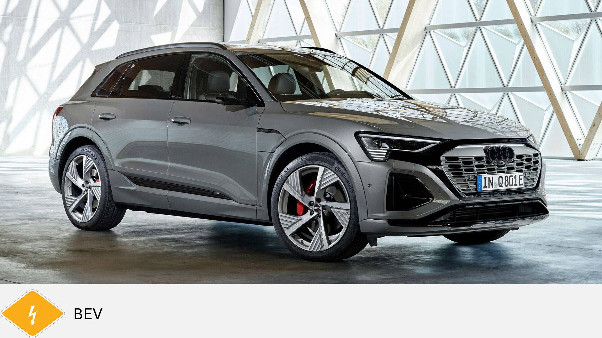 Gray Audi Q8 e-tron SUV stands in light-flooded room with glass structure. Yellow BEV symbol with lightning bolt below.