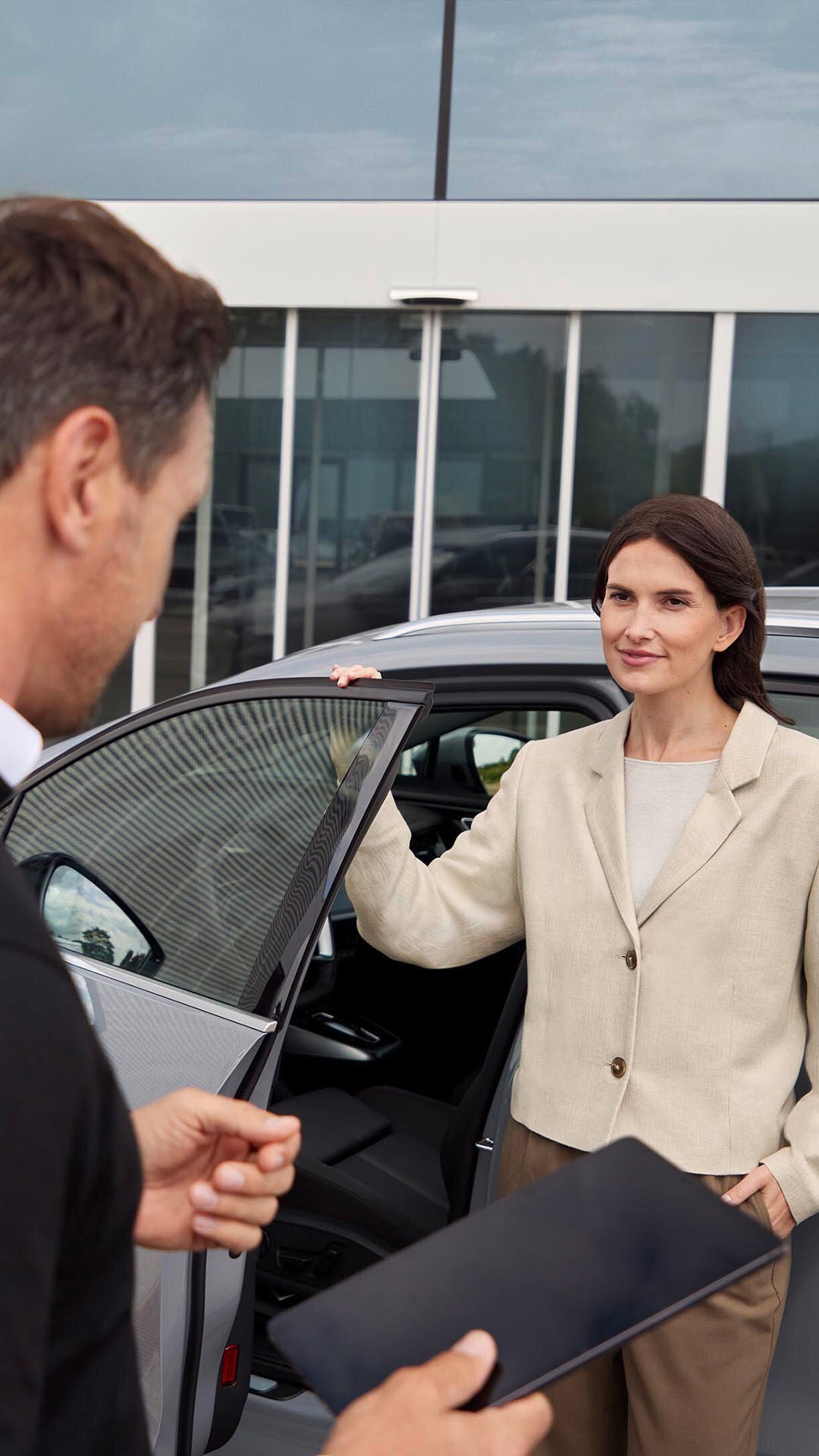 Frau steht an geöffneter Autotür vor Audi-Gebäude, Mann mit Tablet im Vordergrund, moderne Glasfassade im Hintergrund.
