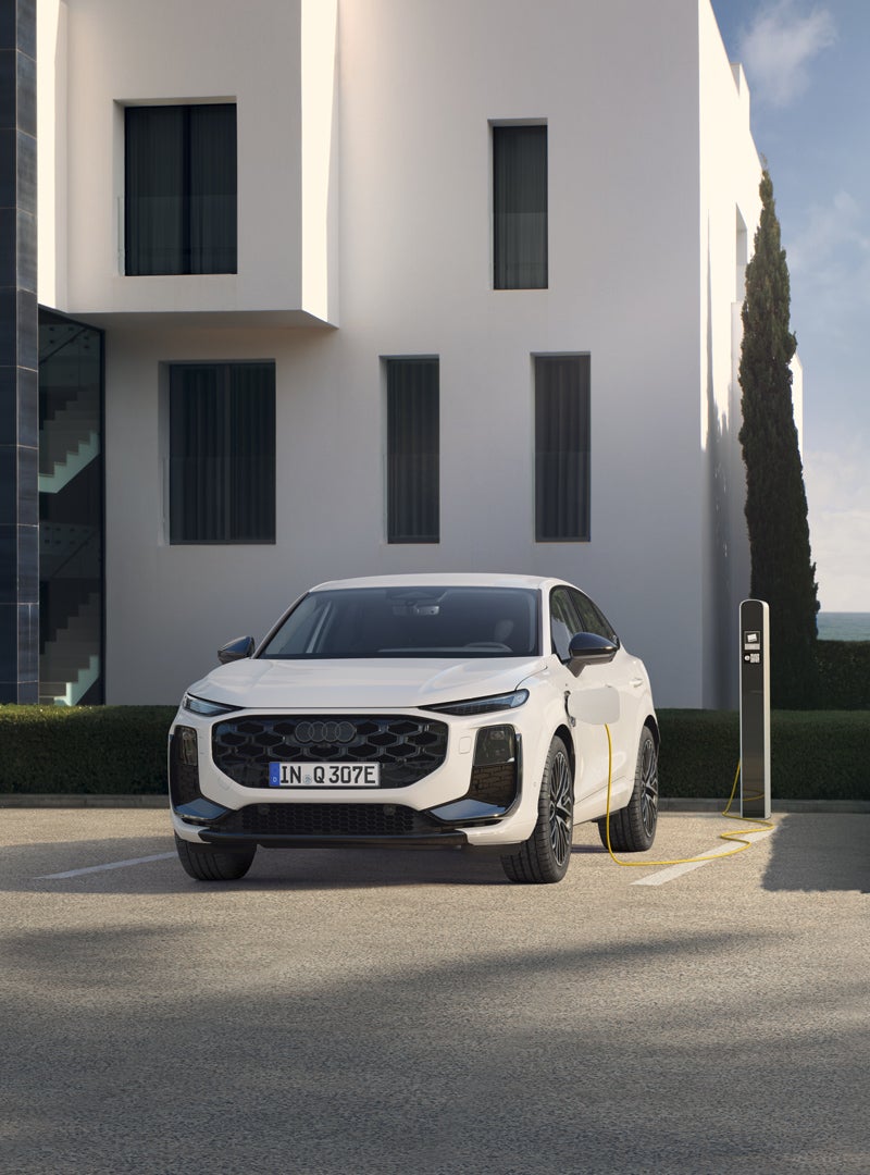 L’immagine raffigura l’Audi Q3 Sportback e-hybrid. Di fronte a un edificio residenziale con una colonnina di ricarica in fase di ricarica.