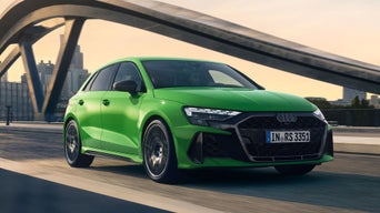 Hellgrüner Audi RS 3 Sportback fährt über Brücke bei Sonnenuntergang, moderne Architektur im Hintergrund.