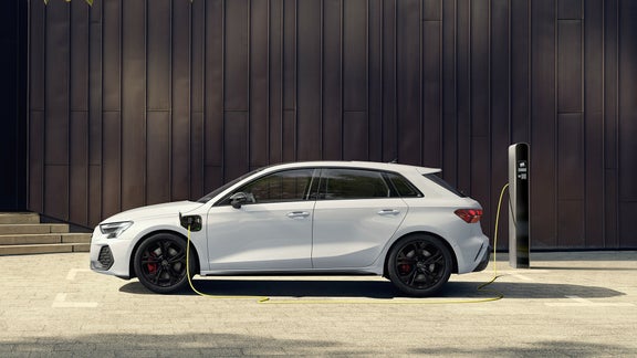 Ein weißer Audi A3 Sportback TFSI e steht seitlich an einer Ladestation, während das gelbe Ladekabel angeschlossen ist. Im Hintergrund eine moderne dunkle Metallfassade, mit einem Baum, dessen Blätter in die Szene ragen.