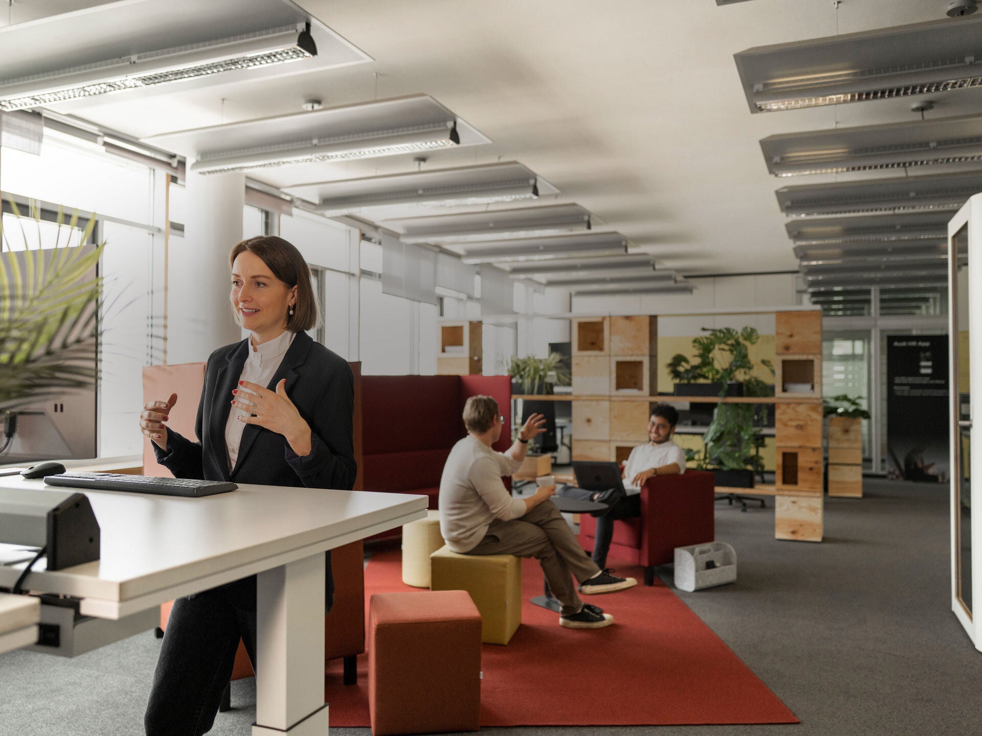 Three people are in an office, two are sitting and talking and a woman is standing at a desk talking into a computer screen