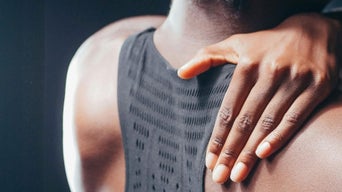 Hand liegt auf Schulter einer Person in ärmellosem Oberteil mit Textmuster, dunkle Stimmung, gedämpftes Licht.