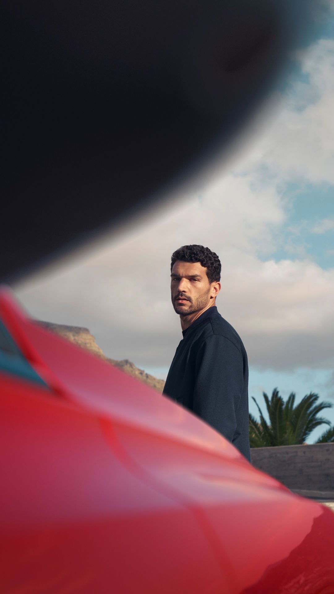 A person in a black jacket stands by a red car, with palm trees and a cloudy blue sky in the background.