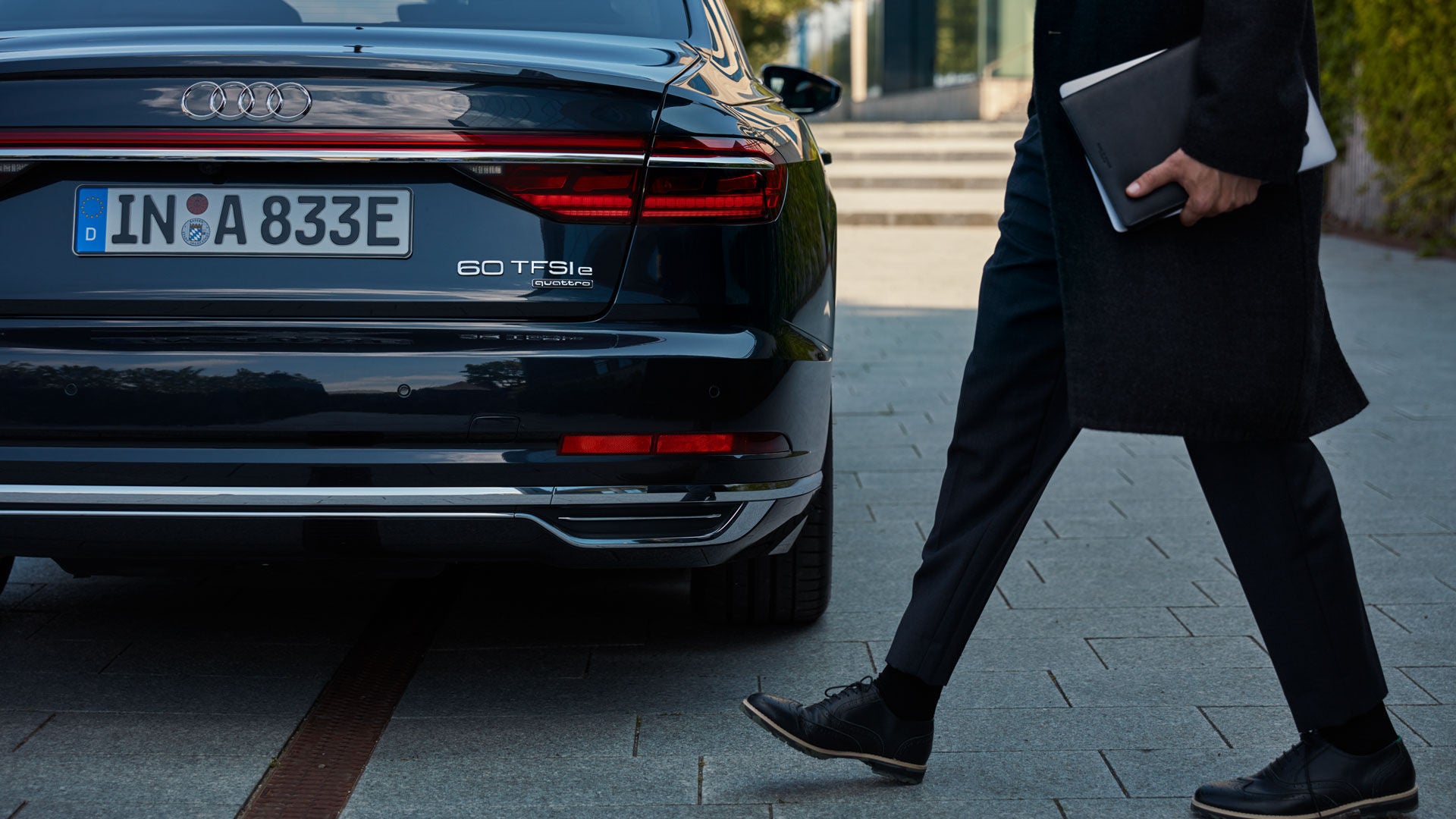 Ein Mann mit Laptop läuft an einem schwarzen Audi A8 60 TFSI e vorbei, Heckansicht des Fahrzeugs im Fokus.