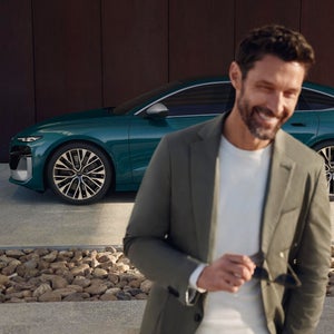 A smiling man in a green blazer holds sunglasses, standing in front of a sleek teal car. The scene exudes a modern and stylish ambiance.