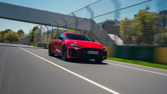 Ein roter Audi RS 5 Avant fährt über die Rennstrecke im Audi driving center Neuburg. Im Hintergrund ist eine Fussgängerbrücke zu sehen. Rechts ist ein Absperrzaun zu erkennen.