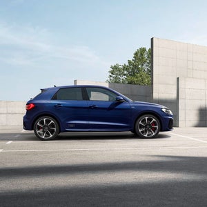 A blue Audi A1 is parked outdoors against a concrete wall, with green foliage in the background. The sky is clear, evoking a calm and modern vibe.