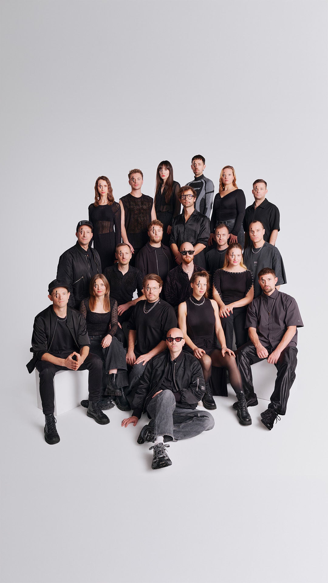 Große Musikgruppe in schwarzer Kleidung posiert im Studio vor hellem Hintergrund; mehrere Reihen, sitzend und stehend.