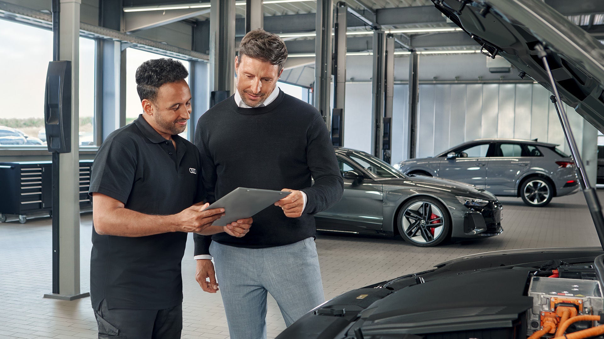 Zwei Männer stehen in einer Werkstatt vor einem Audi Modell mit offener Motorhaube und schauen gemeinsam auf den Screen eines Tablets.