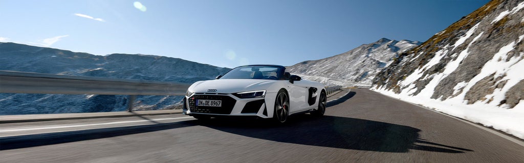 Weißer Audi R8 Spyder V10 performance quattro fährt mit offenem Verdeck bei Sonnenschein durch verschneite Alpenlandschaft auf einer kurvigen Bergstraße.