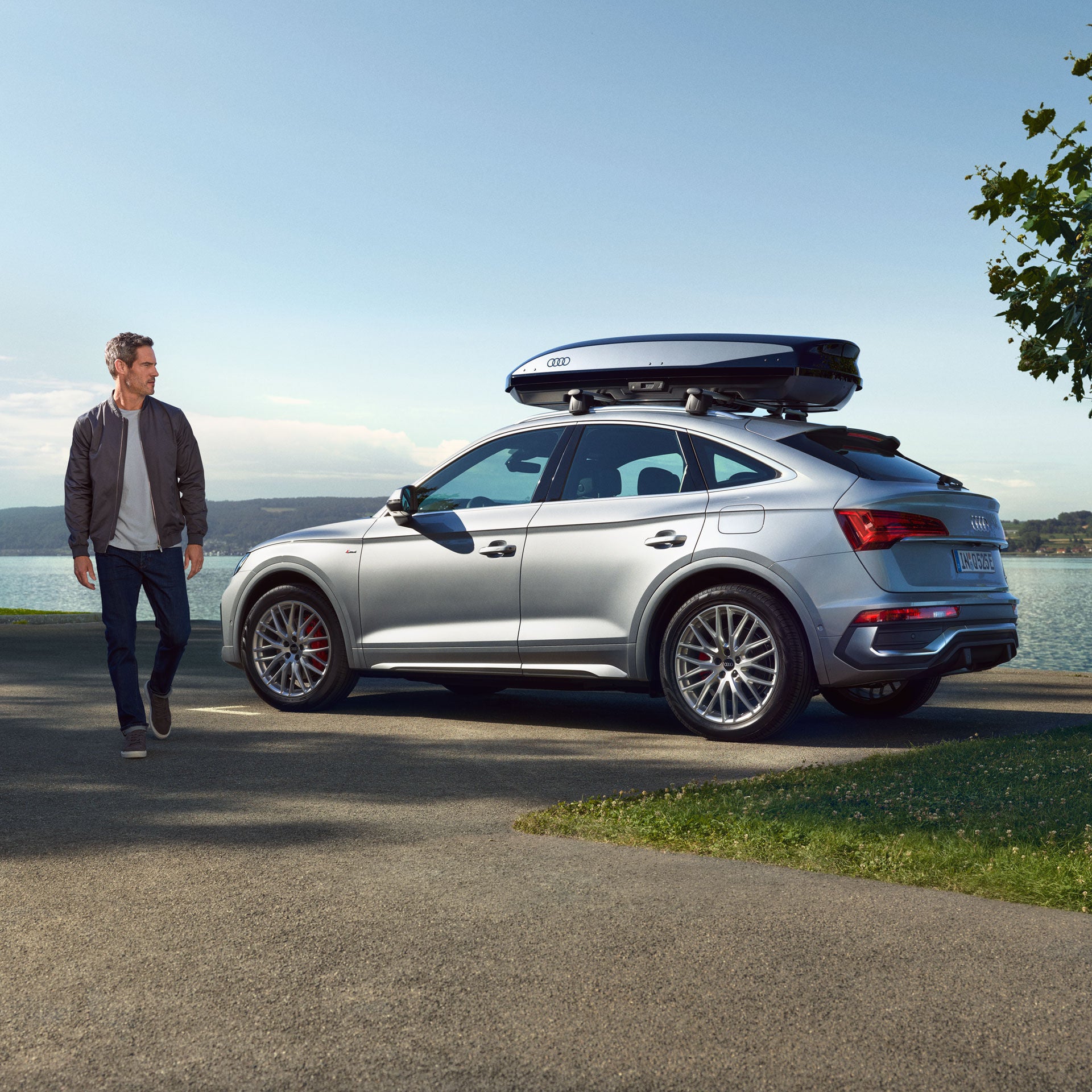 Ein silberner Audi mit Dachbox steht auf einem Parkplatz in Seenähe, während ein Mann in Freizeitkleidung am Fahrzeug vorbeigeht.
