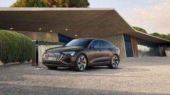 Ein brauner Audi Q8 e-tron steht an einer Ladestation vor einem modernen Gebäude mit schrägem Dach und großer Glasfassade.
