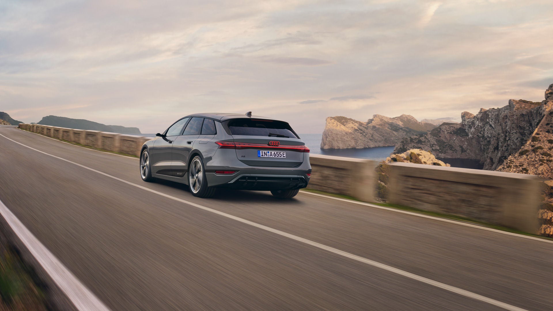 Ein grauer Audi A6 Avant fährt auf einer kurvigen Küstenstraße mit beeindruckender Felslandschaft und Meerblick im Hintergrund. Die Szenerie wird von einer warmen Abendstimmung begleitet.