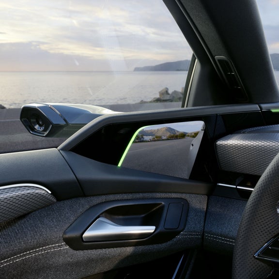 Detailansicht eines digitalen Außenspiegels in einem Audi. Die Kamera auf der Fahrzeugaußenseite überträgt das Bild auf einen Bildschirm im Innenraum. Im Hintergrund ist eine Küstenstraße mit Blick auf das Meer und die Berge zu sehen.