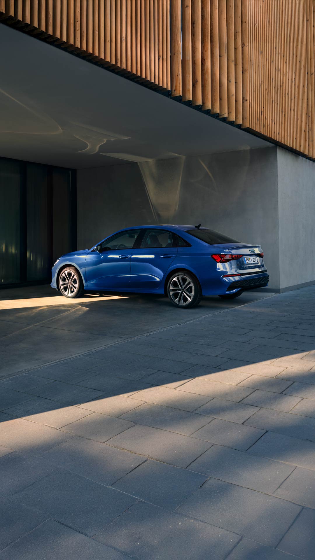 Blaue Audi A3 Limousine steht halb im Schatten vor einer modernen, grauen Gebäudewand.