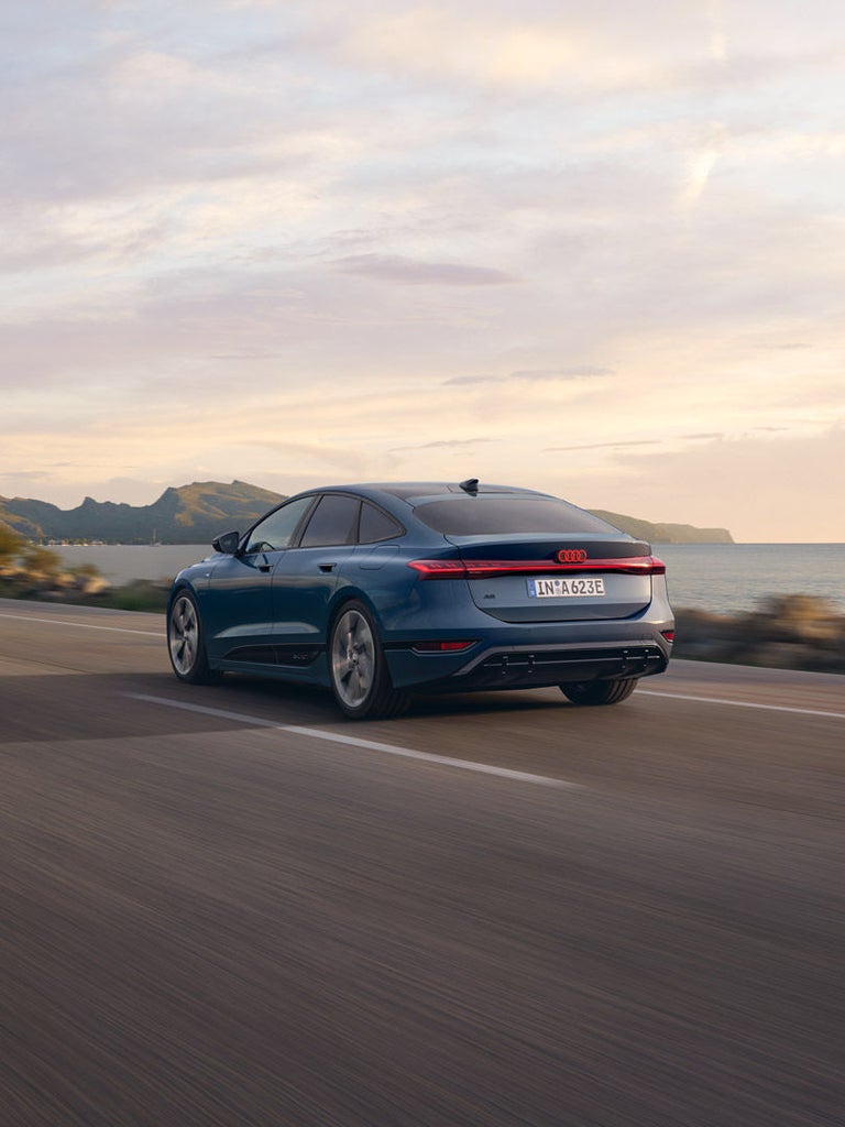 Blauer Audi A6 Sportback e-tron fährt auf Küstenstraße bei Sonnenuntergang, Meer und Berge im Hintergrund.
