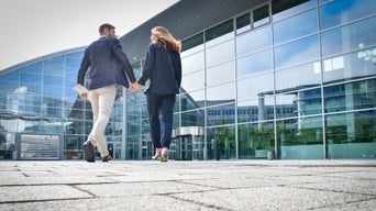 Zwei Personen laufen Hand in Hand auf ein Glasgebäude zu