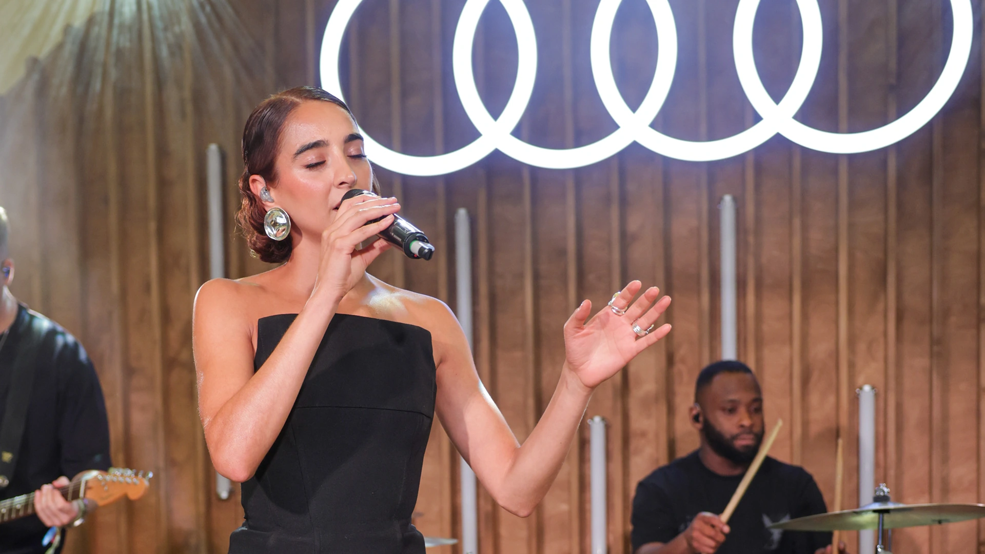 Two performers sing into microphones on stage as a crowd films and cheers, lit by warm stage lighting under a curtained backdrop. Two performers sing into microphones on stage as a crowd films and cheers, lit by warm stage lighting under a curtained backdrop.