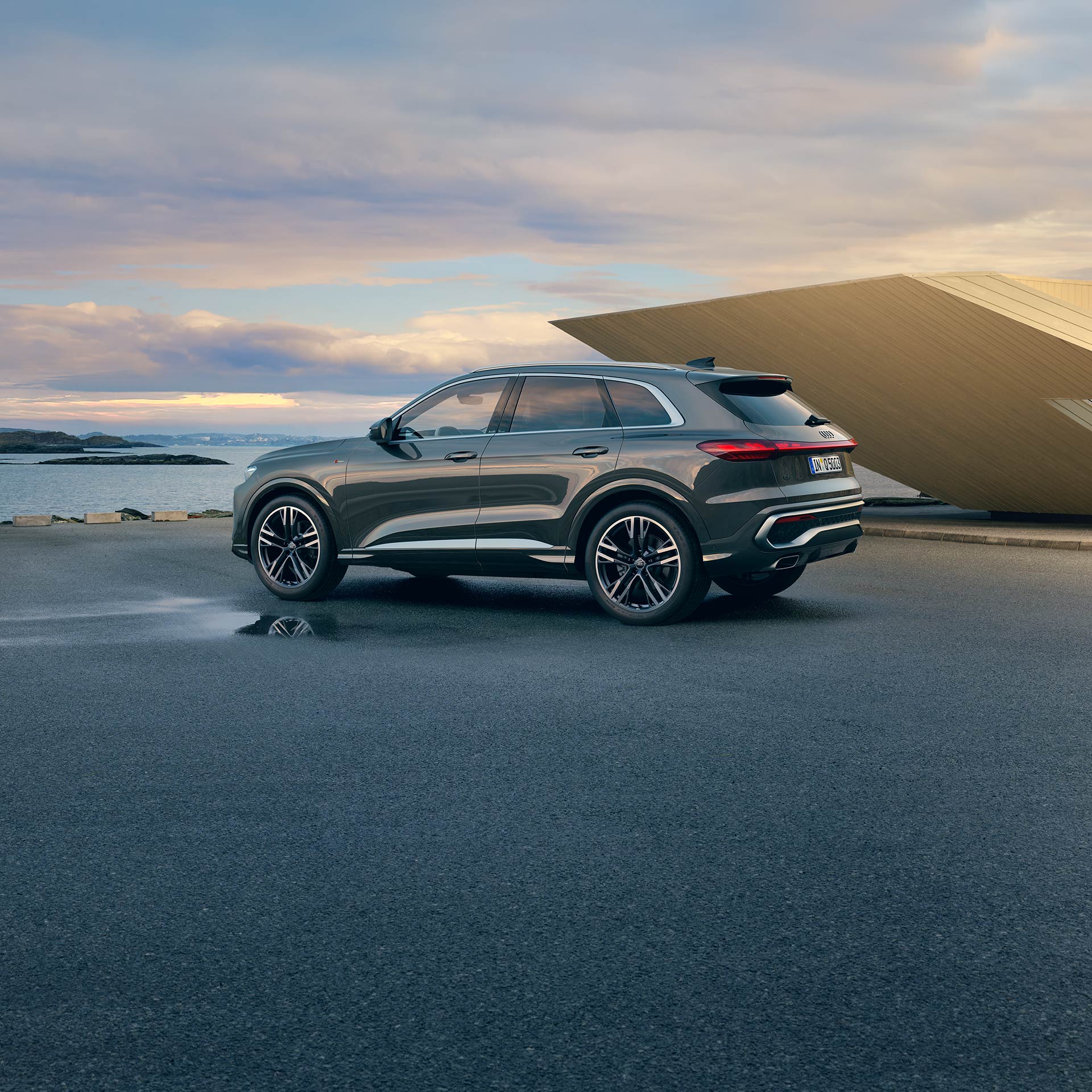 Ein grauer Audi Q5 SUV steht auf einer Parkfläche mit Meerblick und moderner Architektur im Hintergrund.