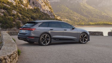 The A6 Avant e-tron is parked sideways on a gravel car park in front of a lake. Tree-covered mountains can be seen in the background.