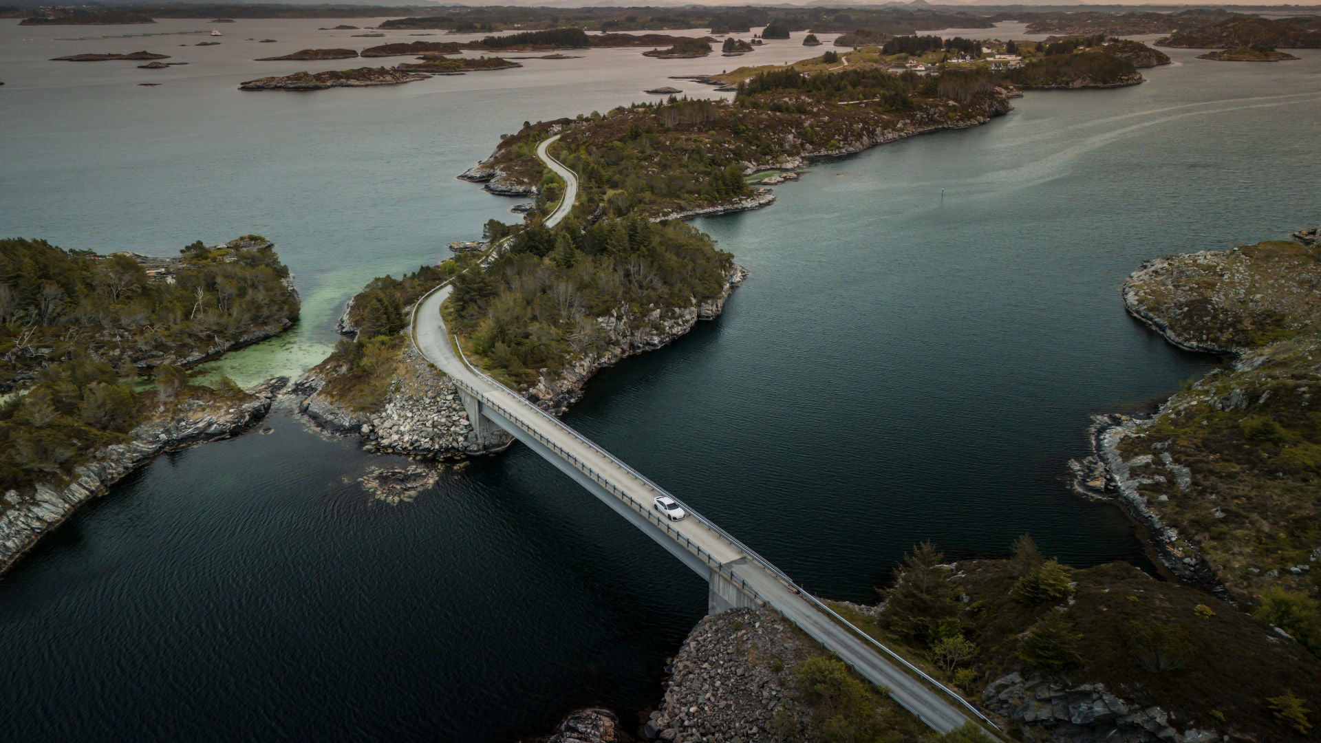 Ein Audi A6 Sportback e-tron in Gletscherweiß Metallic überquert eine Brücke in der zerklüfteten Küstenlandschaft Norwegens. Im Hintergrund befinden sich zahlreiche kleine Inseln. 