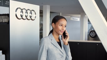 Frau im grauen Blazer telefoniert in einem Audi-Autohaus neben einem Audi-Logo.
