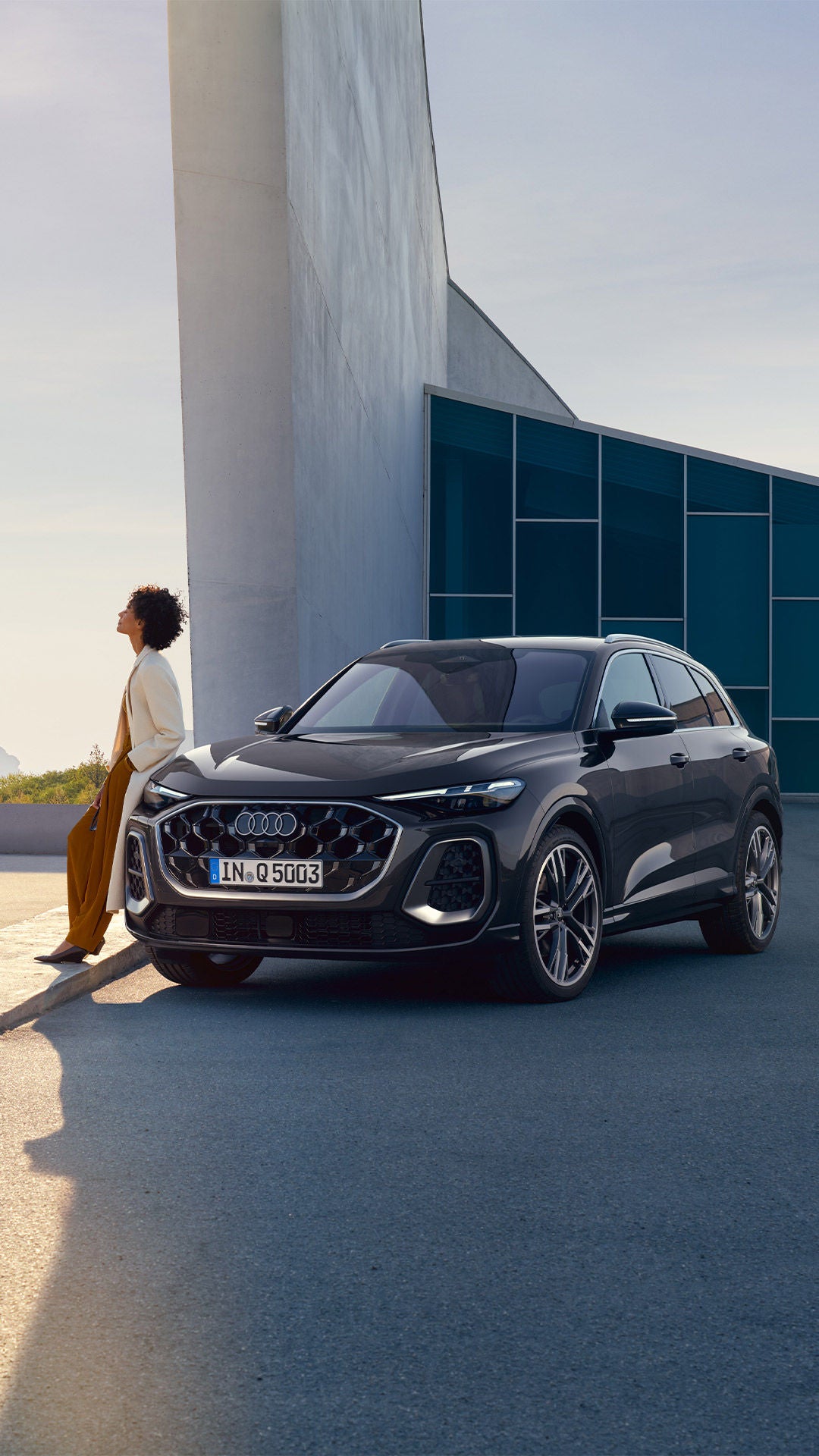 Un SUV Audi moderne de couleur gris foncé garé devant un bâtiment au design architectural contemporain, avec une personne adossée à un muret en arrière-plan.