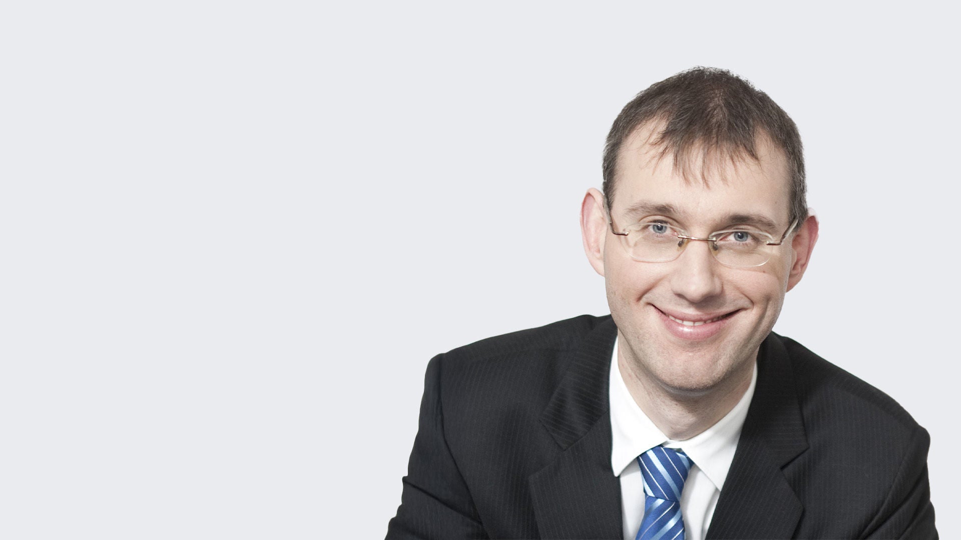 Dr. Steffen Bauer: Porträt eines lächelnden Mann mit Brille, schwarzem Anzug und weißem Hemd und blau, gestreifter Krawatte vor grauem Hintergrund.