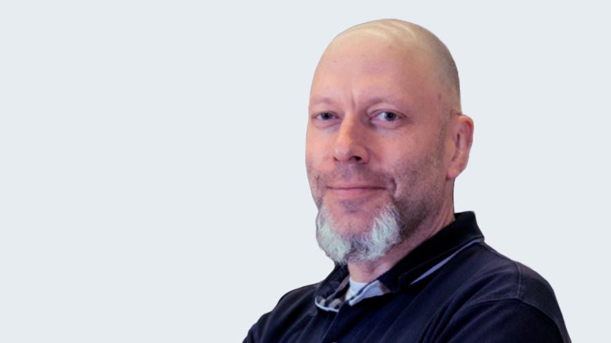 Udo Frittrang: Porträt älterer, lächelnder Mann mit längerem grauen Kinnbart vor grauem Hintergrund.