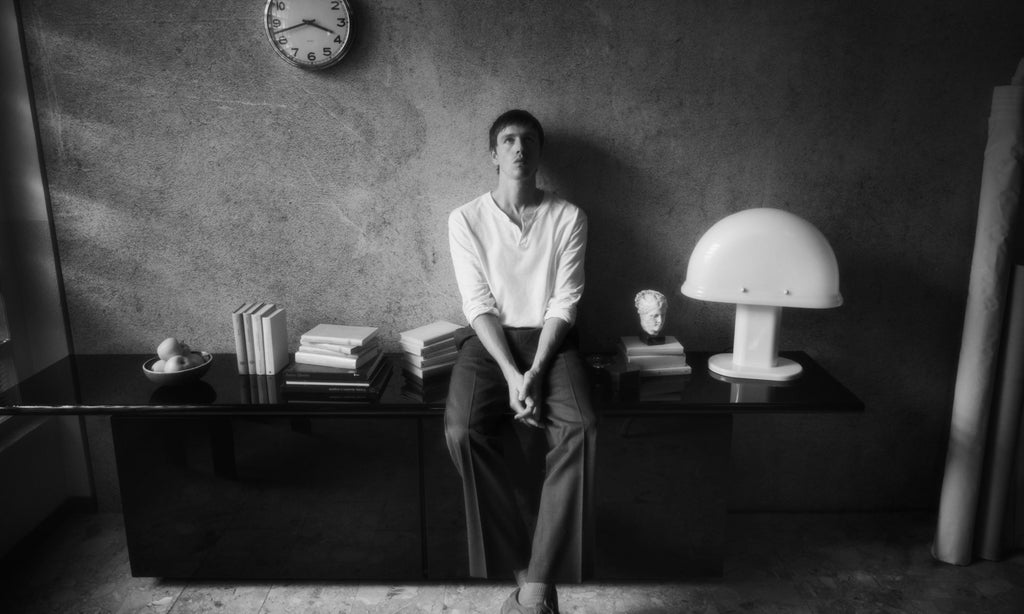 Harris Dickinson is sitting on a desk, deep in thought.