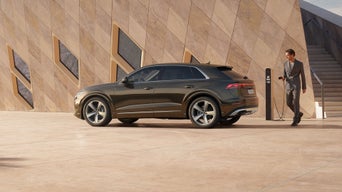 Der Q8 SUV e-hybrid von der Seite, vor einem futuristischen Gebäude parkend. Ein modisch gekleideter junger Mann steht hinter dem Fahrzeug und hält den Stecker einer Ladesäule in der Hand.
