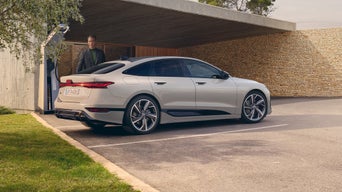 Ein heller Audi A6 Sportback e-tron wird vor einem modernen Haus an einer Ladestation geladen, ein Mann steht daneben.
