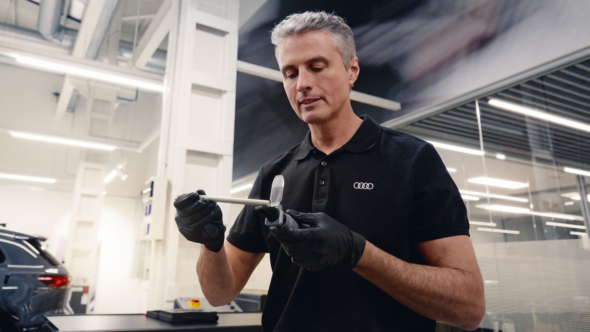 Audi Mitarbeiter in schwarzem Poloshirt hält in der Werkstatt ein Messgerät und prüft eine Fahrzeugkomponente.