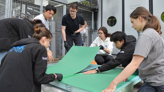 A few people sit and stand around a green cardboard on the floor
