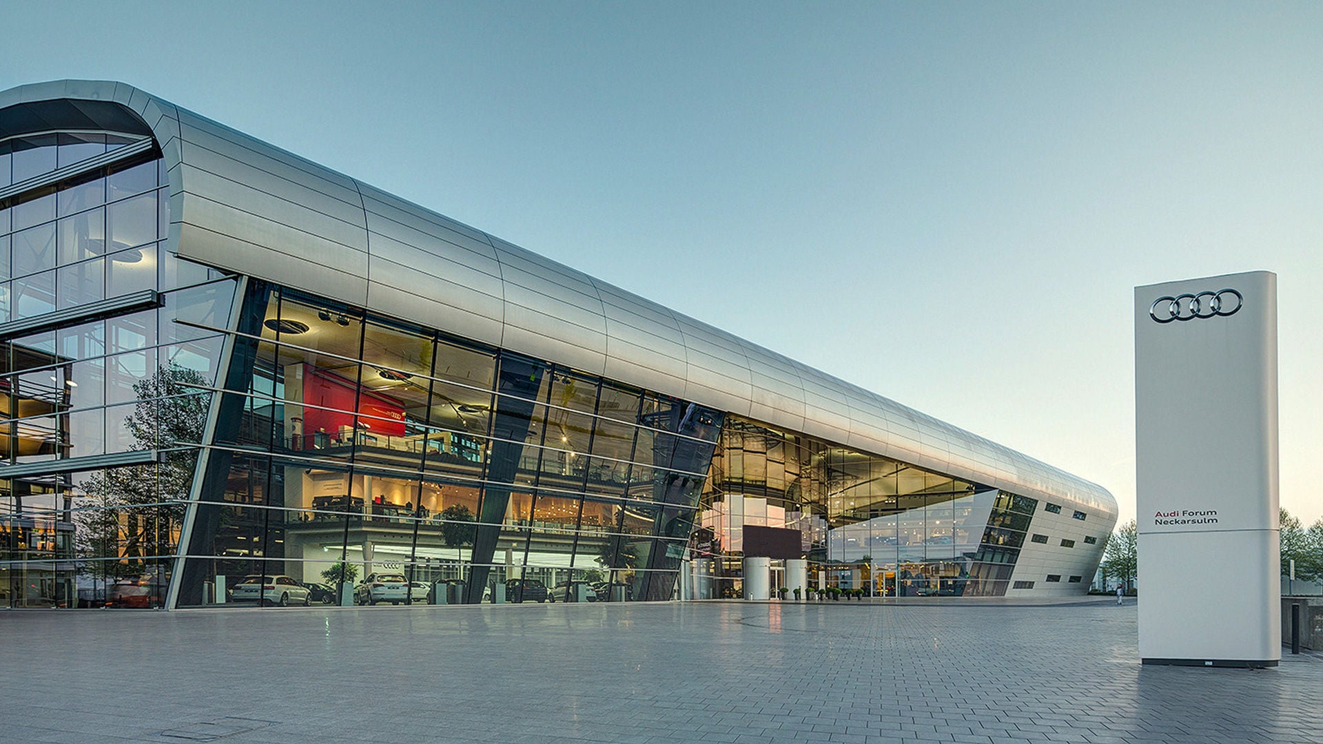 Moderne Glasfassade des Audi Forum Neckarsulm bei Tageslicht mit großem Audi-Logo im Vordergrund.