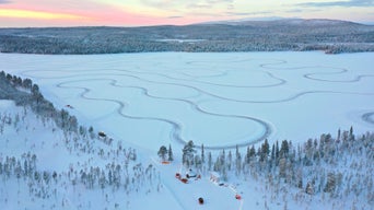 A snow-covered landscape with winding tracks on a frozen lake, surrounded by woodland and a wintry sky.