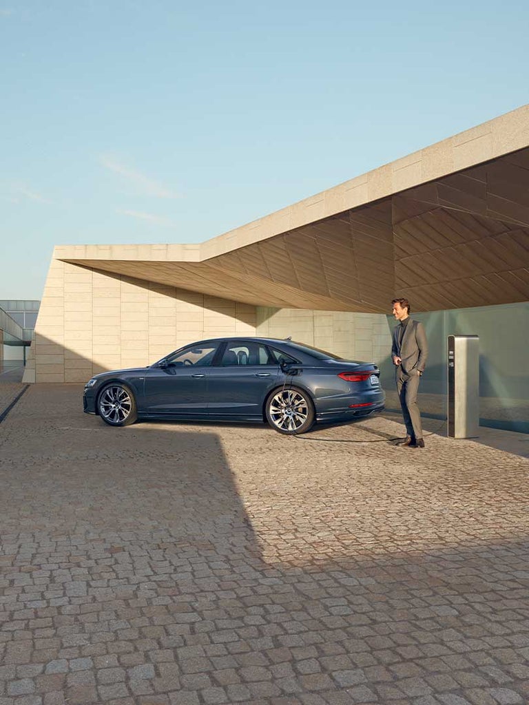 Ein dunkelblauer Audi A8 60 TFSI e an Ladesäule im Sonnenschein, Mann in grauem Anzug steht daneben. Im Hintergrund ein modernes, beiges Gebäude.
